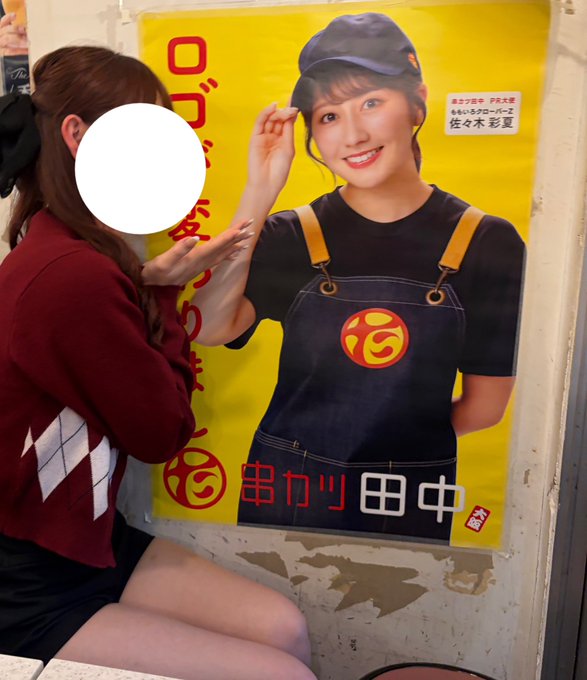 A person in a red argyle sweater poses playfully next to a large yellow promotional poster featuring a woman in denim overalls, with Japanese text including "串カツ田中" (Kushikatsu Tanaka) visible.