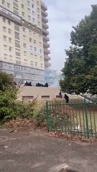 An urban scene showing what appears to be a confrontation near a tall residential building, with figures visible on a low rooftop or wall behind a green metal fence.