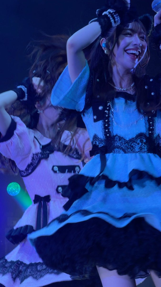 Two smiling performers in elaborate blue and pink frilly costumes with black lace trim perform on stage, one holding a microphone while dancing.
