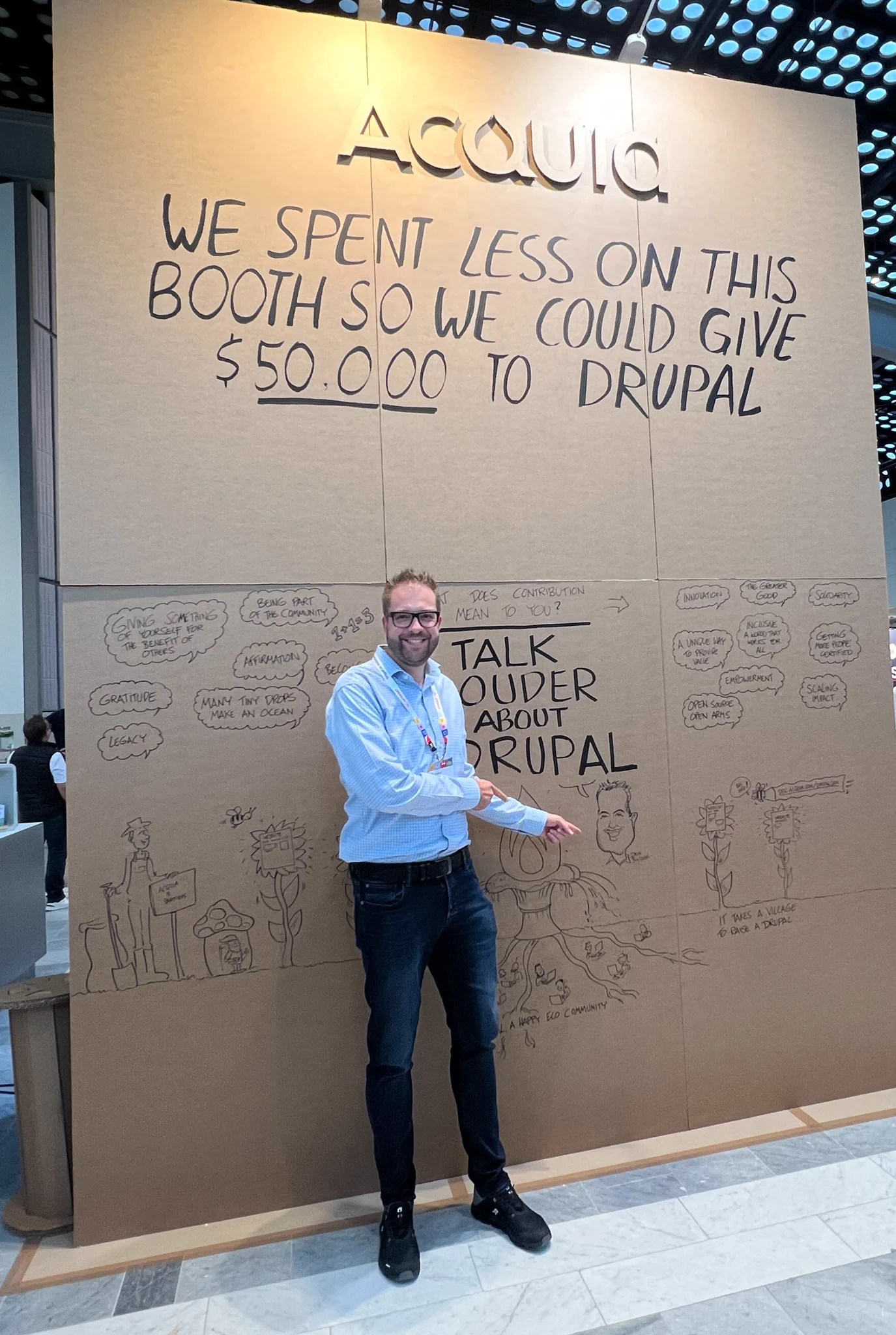 Man standing in front of a large cardboard booth with handwritten text about donating $50,000 to Drupal from Acquia.