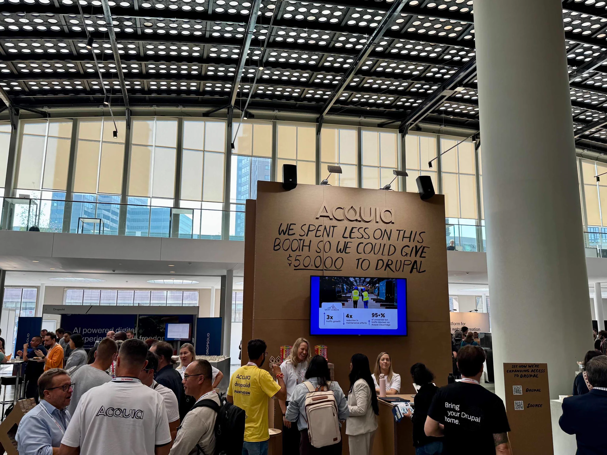 Crowded conference booth with a sign stating savings were donated $50,000 to Drupal, featuring a digital screen and attendees interacting.