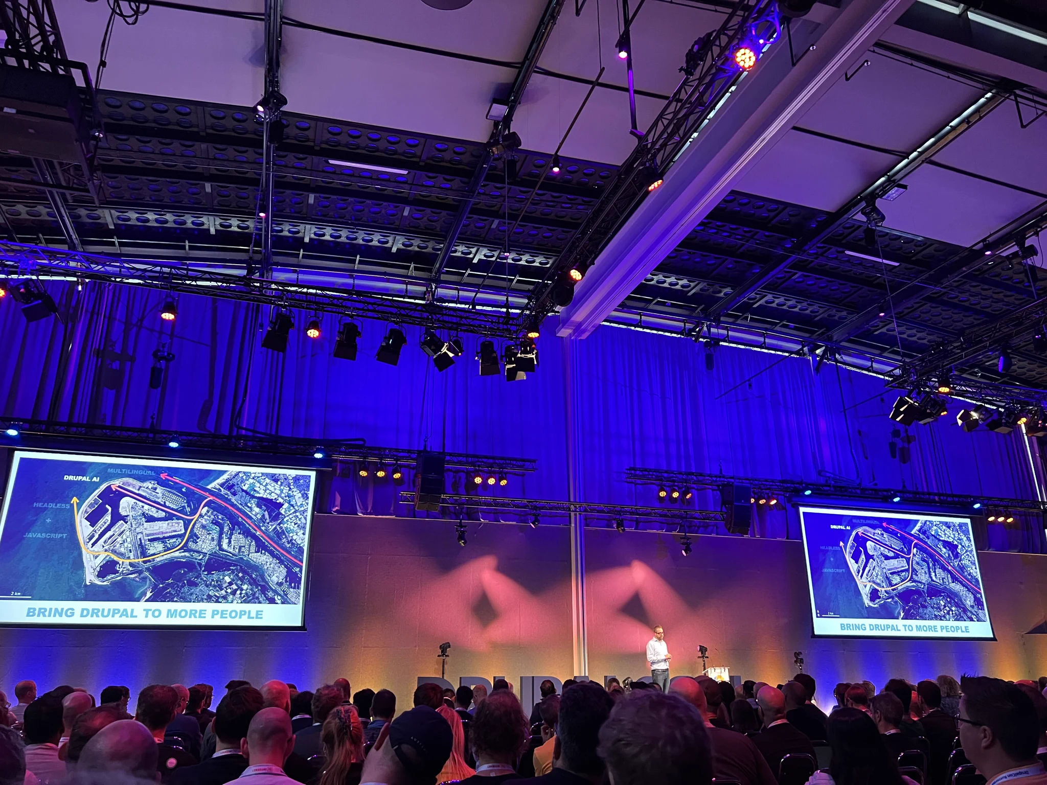 Crowd attending a tech conference with two large screens displaying a map and the slogan 'Bring Drupal to more people'.