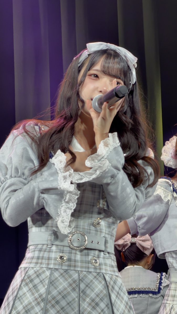A Japanese idol performing on stage wearing a gray plaid dress with lace details and a large bow headband, holding a microphone.
