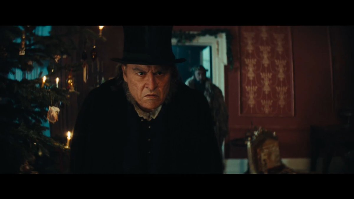 Johnny Depp portrays Ebenezer Scrooge in a dark, period setting, wearing a black top hat and Victorian coat, standing in a dimly lit room with a decorated Christmas tree and another figure in the background. The image aligns with the promotional content for the film "Ebenezer," highlighting the movie's moody, gothic atmosphere as Scrooge.