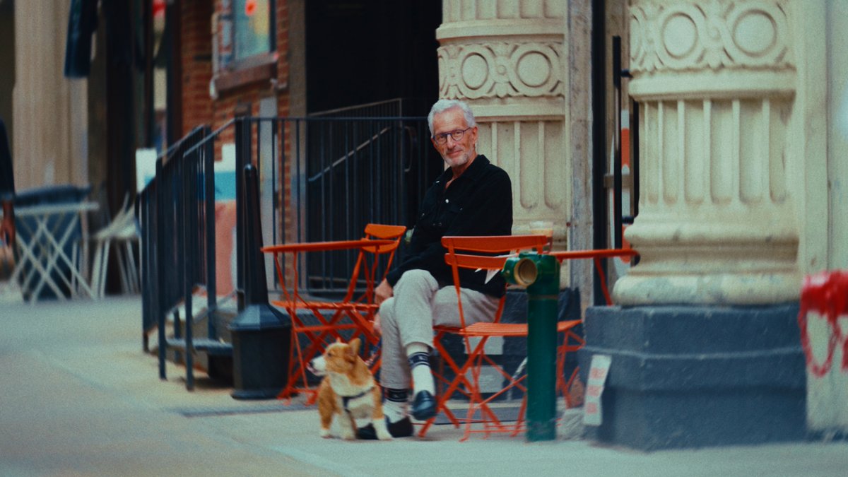 A gray-haired man wearing glasses sits at an outdoor cafe next to a corgi dog on a sidewalk in front of an ornate building.