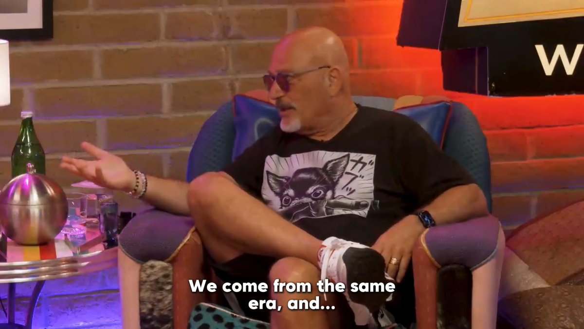 A bald man wearing sunglasses and a black t-shirt featuring a chihuahua graphic with Japanese text sits in a teal armchair with the subtitle "We come from the same era, and..." displayed below. The setting includes a brick wall backdrop with colorful lighting, a small side table holding a green bottle and metallic sphere, consistent with the "Club Random" podcast set referenced in the caption.