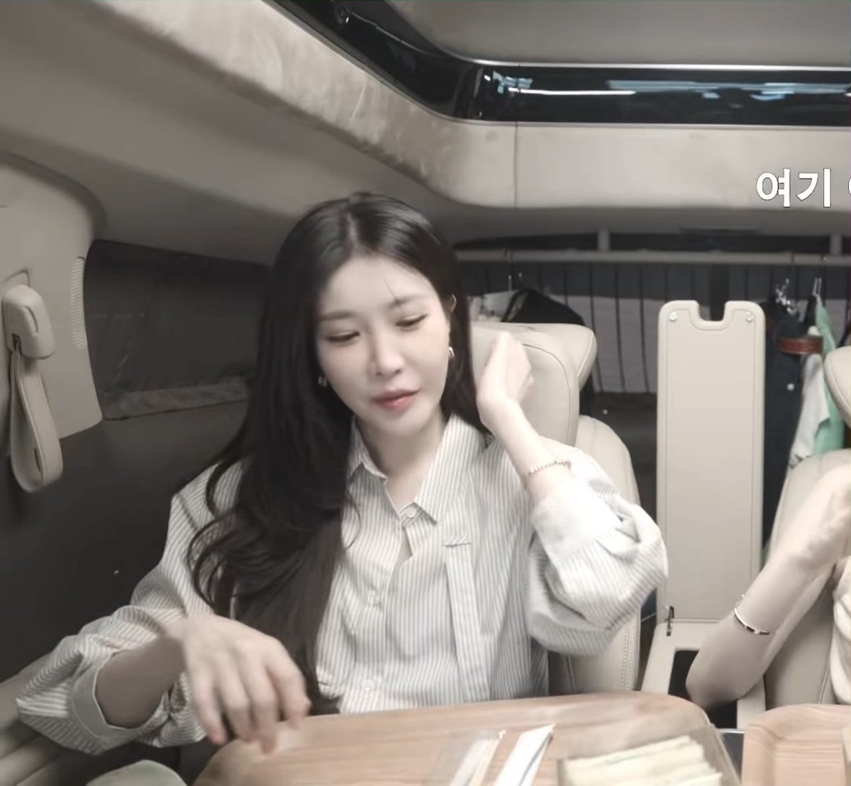 A woman with long dark hair wearing a striped blouse is seated inside what appears to be a vehicle or studio setting, with Korean text "여기" visible in the upper right corner. The caption expresses admiration for her beauty, fitting the image of her looking down with a soft, composed expression.