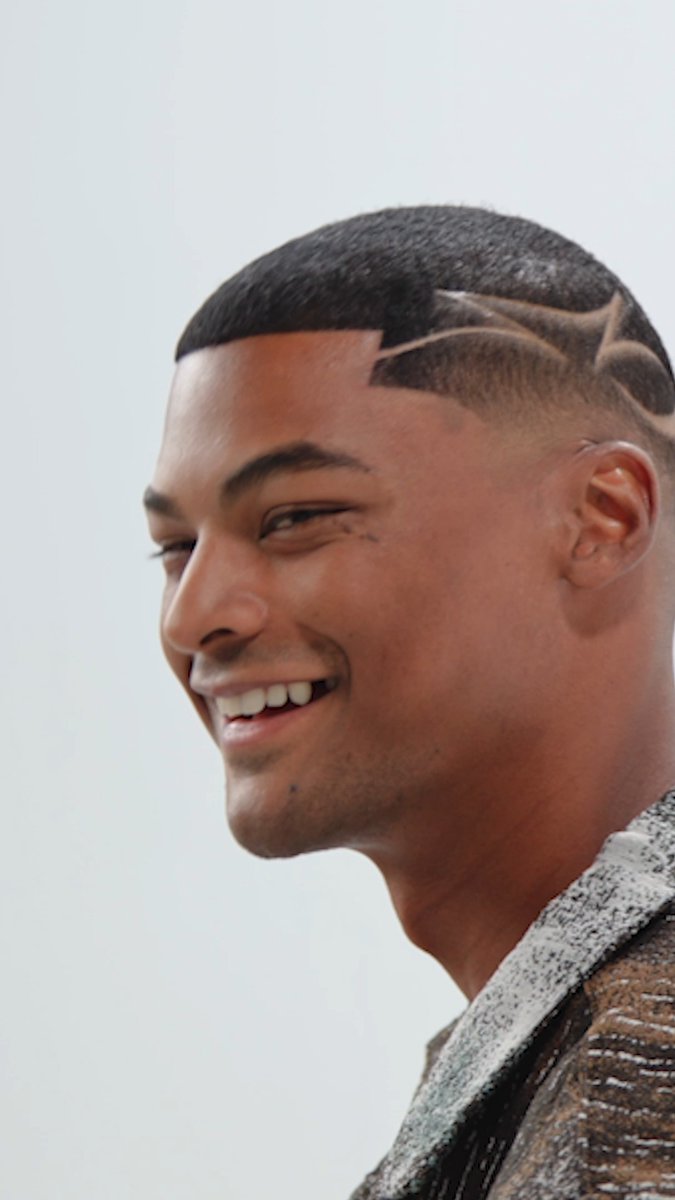A smiling man with a distinctive haircut featuring shaved designs on the side of his head, wearing a grey textured jacket.