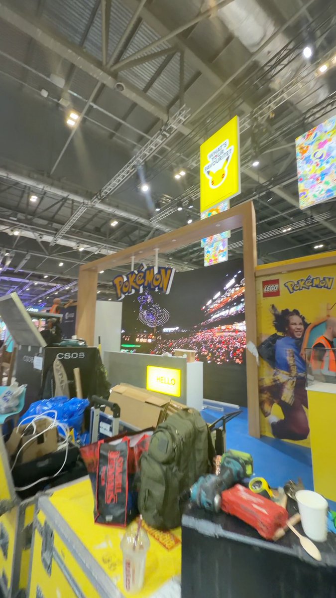 A behind-the-scenes view of a Pokémon and LEGO booth at a convention or trade show, featuring display screens, colorful signage, and various equipment like a backpack and power tools in the foreground.
