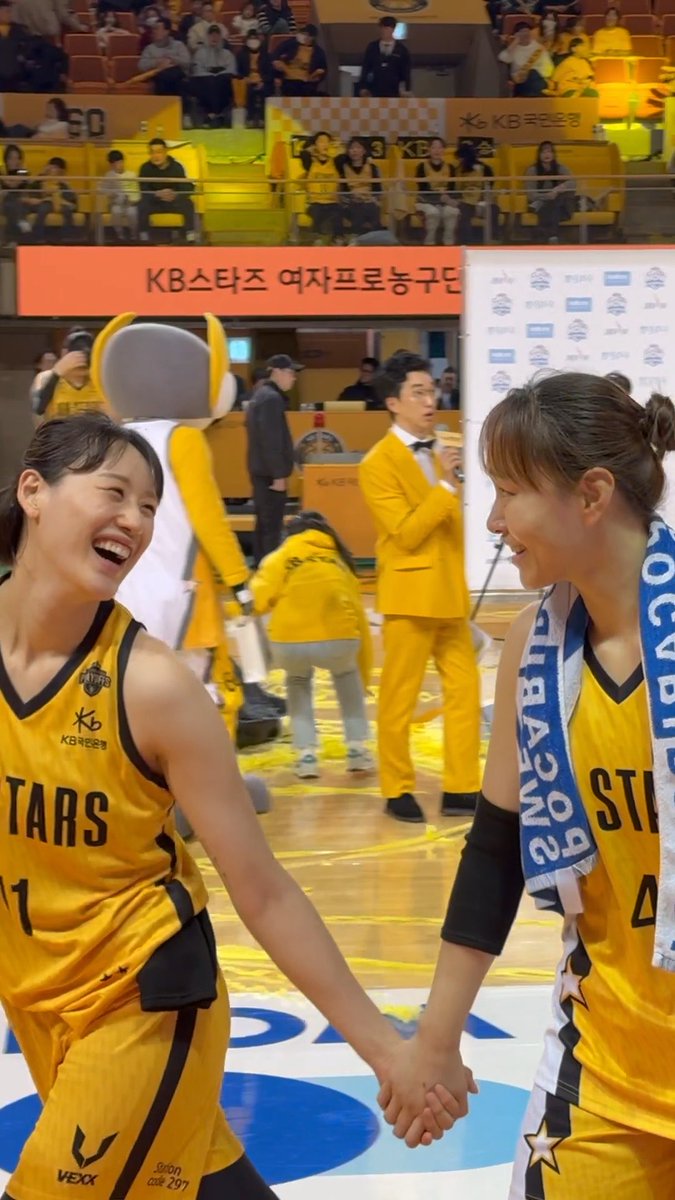 Two female basketball players in yellow 'STARS' uniforms smile and hold hands on the court, with Korean signage for 'KB스타즈' (KB Stars) and a yellow mascot visible in the background.