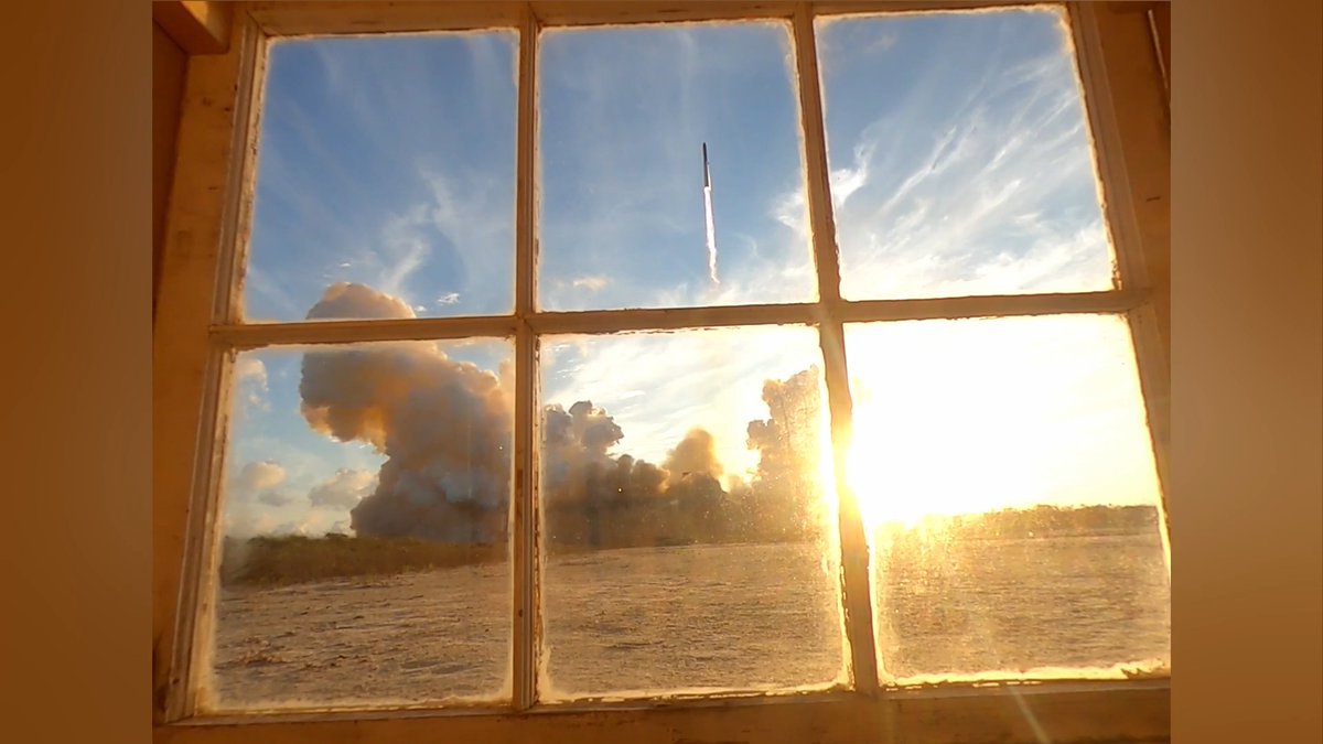 A rocket launches skyward, leaving a trail of white smoke against a blue sky, viewed through a six-pane wooden window with the golden sun setting over a coastal landscape. The scene captures the spectacle of a rocket launch as seen from a window overlooking the water—perfect for watching both the launch and potential booster catch.
