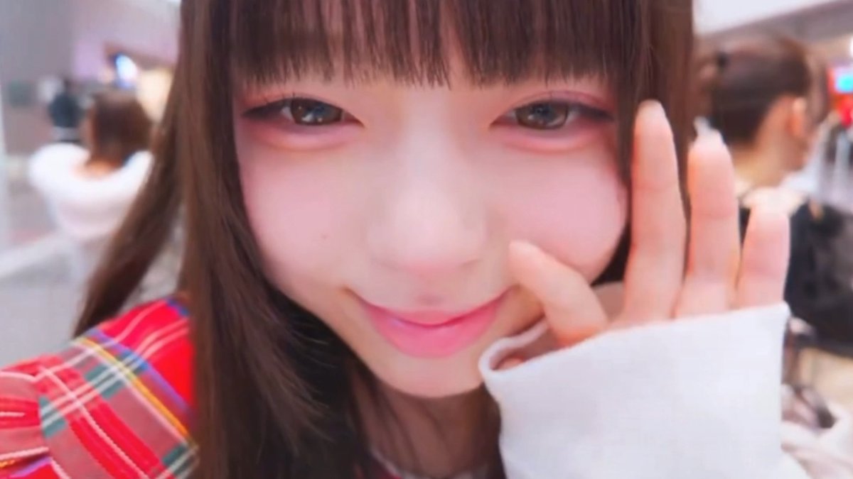 A close-up selfie of a smiling young woman with pink eye makeup and straight bangs, holding her hand near her face and wearing a red plaid garment.
