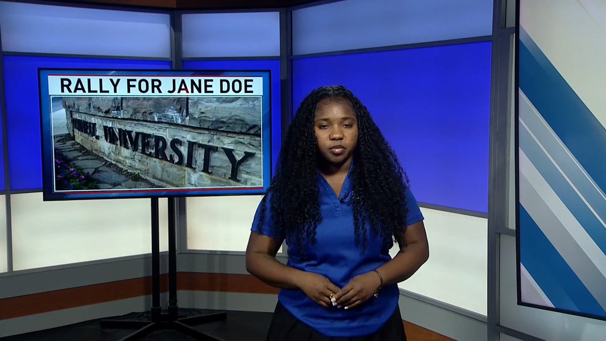 A young Black woman with long curly hair wearing a blue polo shirt stands in a news studio in front of a screen displaying 'RALLY FOR JANE DOE' alongside an image of a 'CORNHELL UNIVERSITY' sign.