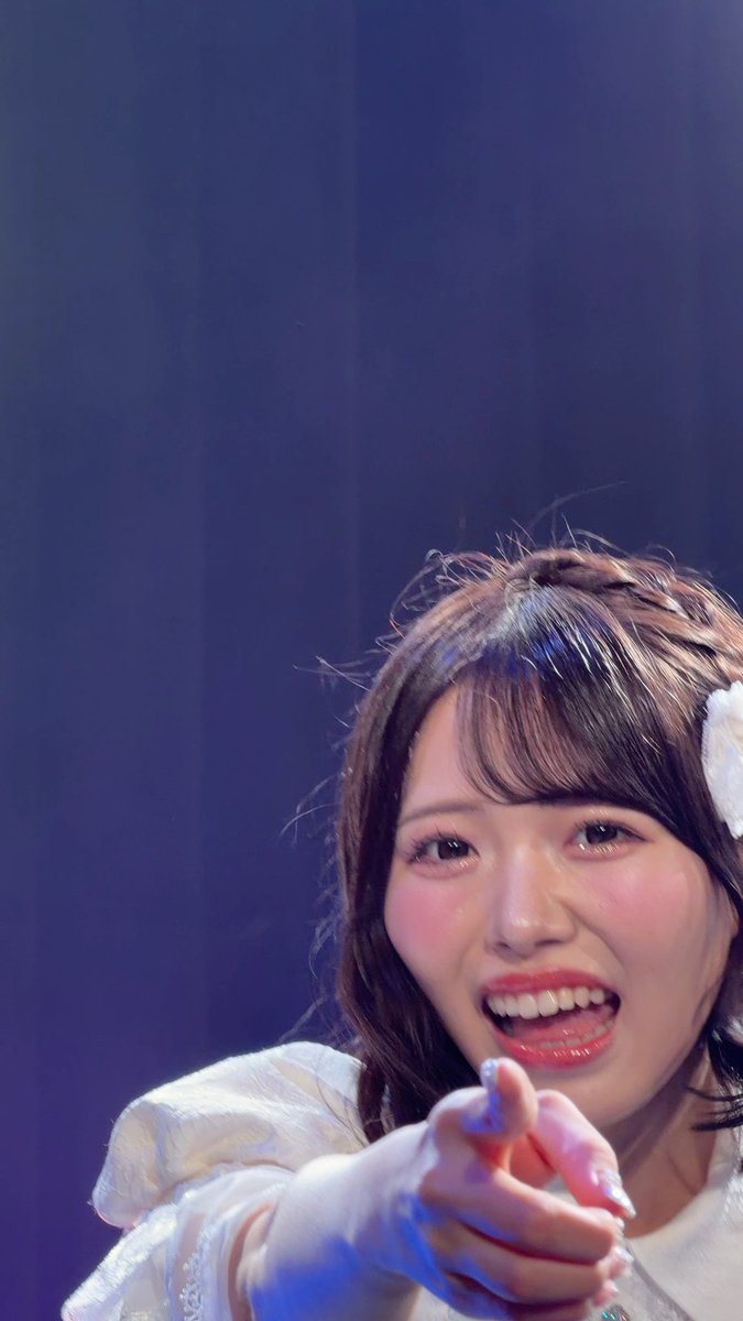 A close-up photo of a Japanese idol/performer's smiling face with brown hair, wearing a white outfit with a hair accessory, pointing toward the camera.