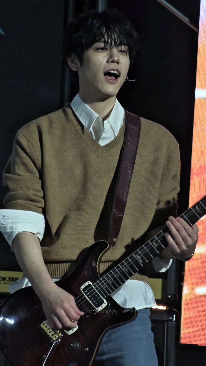 A young man wearing a brown sweater over a white collared shirt plays a dark brown electric guitar on a stage.