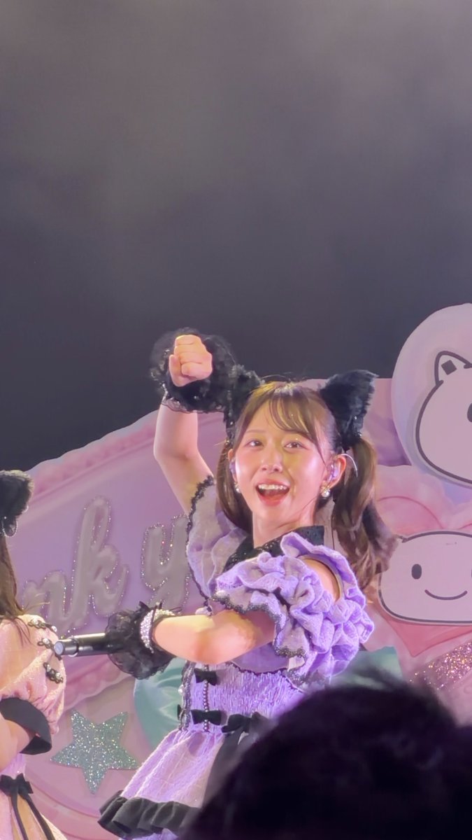 A performer in a purple ruffled dress with black cat ears enthusiastically raises her fist while singing on stage, with a cute cartoon character backdrop visible behind her.