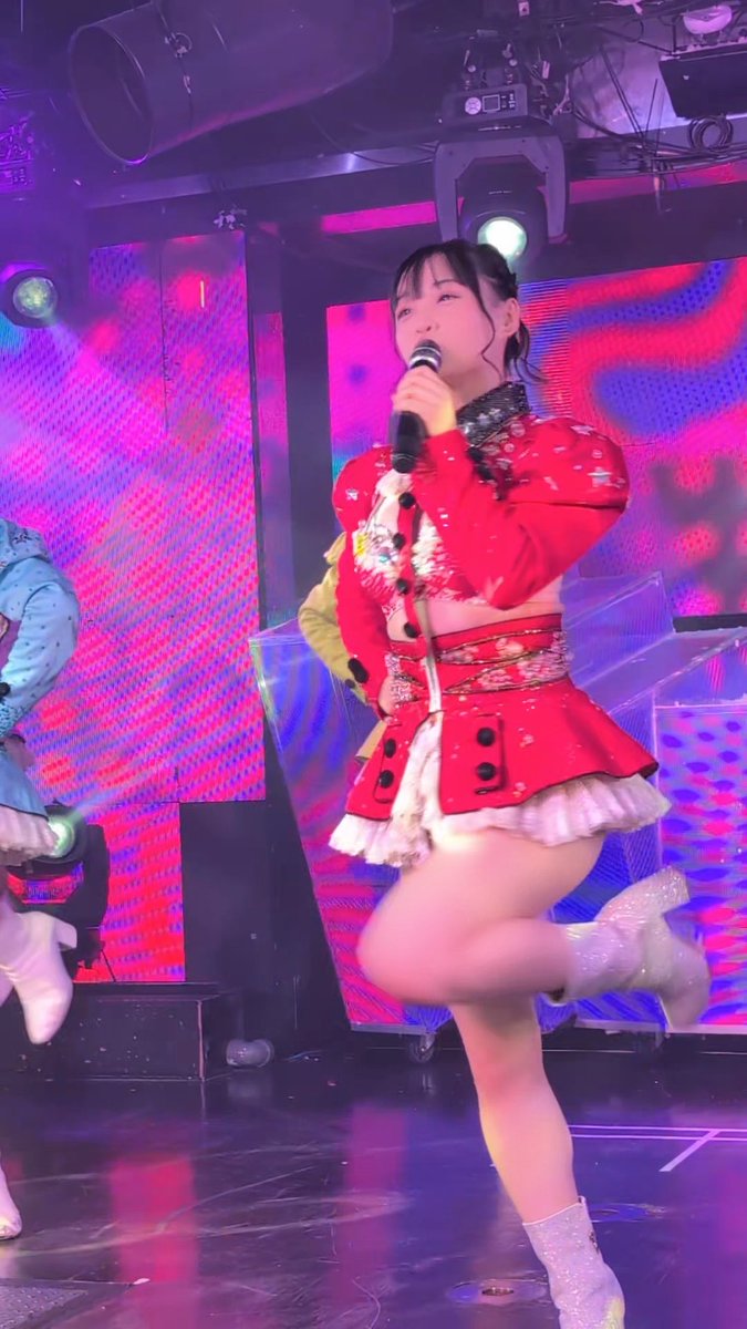 A young woman in a red and white idol-style outfit sings into a microphone while performing on a brightly lit stage with pink and blue lighting.