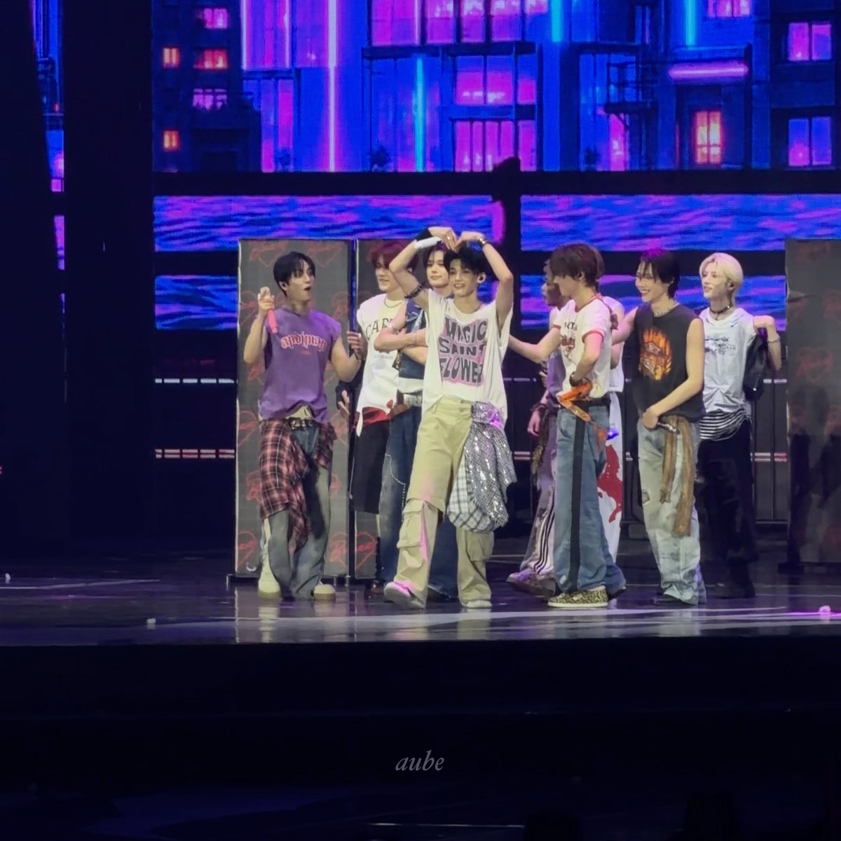 A group of performers on stage pose together with one member forming a heart shape with their arms; the scene features a purple-lit cityscape backdrop and a member wearing a 'MUSIC SAINT FLOWER' shirt, with 'aube' watermarked at the bottom.