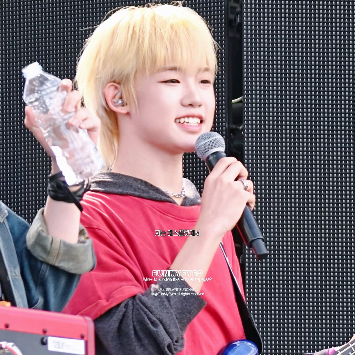 A blonde-haired performer speaking into a microphone at what appears to be a concert, wearing a red shirt and holding a water bottle, with Korean subtitle text and 'EUNMYEYES For SPLUNK EUNCHAN @EunMyEyes' watermark.