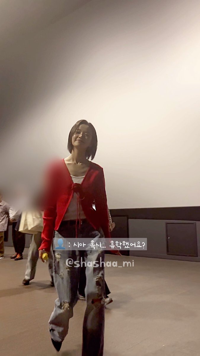 A woman in a red cardigan and distressed jeans stands in an indoor venue with the text overlay ': 사아 혹시... 류학셩여요? @shashaa_mi'.