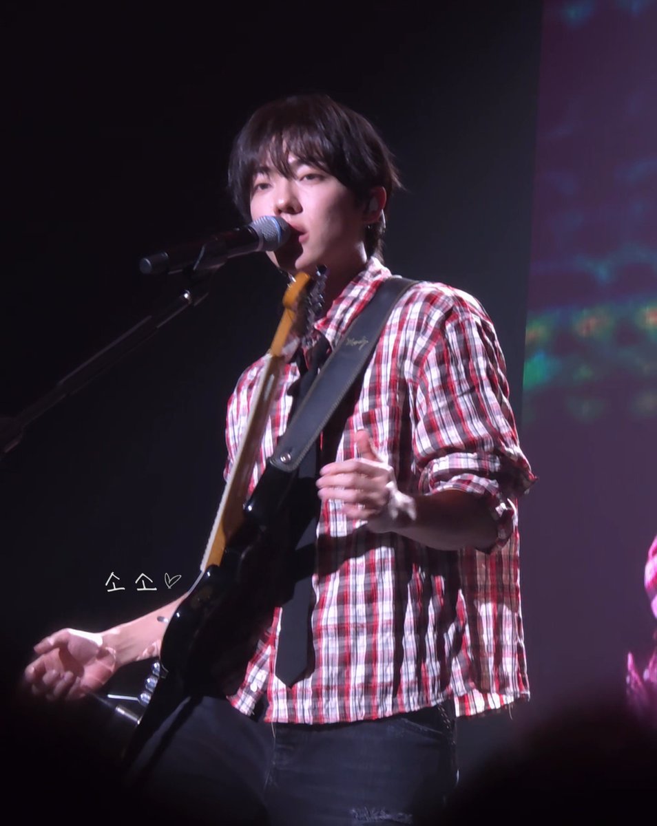 A young man with dark hair performs on stage, singing into a microphone while playing an electric guitar, wearing a red and white plaid shirt and a dark tie.