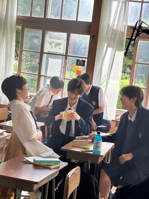 Four young men in Japanese school uniforms laughing and smiling together while sitting at wooden desks in a sunlit classroom with sheer curtains.
