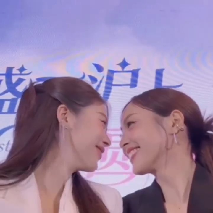 Two women are shown in profile facing each other, smiling intimately, with Chinese characters visible on the backdrop behind them.