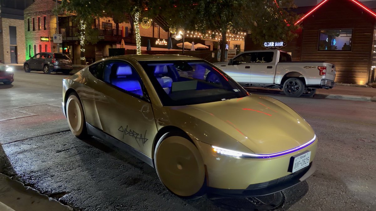 A gold-colored Tesla Cybertruck with 'CYBERTRUCK' written on the door is parked on a city street at night, with blue interior lighting visible through the windows.