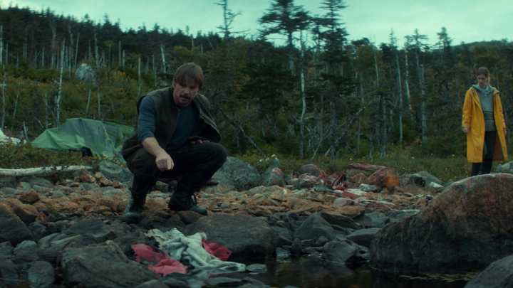 A man crouches on a rocky, desolate shoreline examining debris and scattered clothing, while a woman in a bright yellow raincoat stands watch against a backdrop of windswept, dead trees. The bleak, rugged landscape reflects the ominous tone of the promotional post teasing the Thursday premiere of "BELOW."