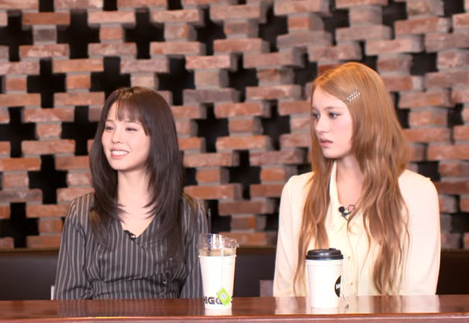 Two women sit at a table during an interview, one with black hair in a striped shirt and one with long brown hair and hair clips.