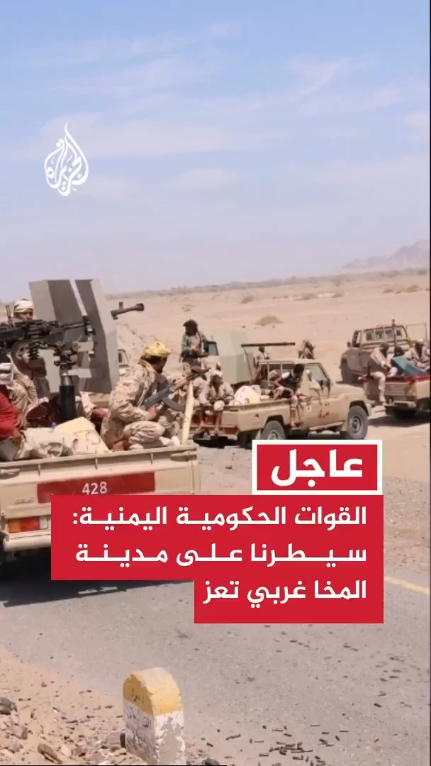 Armed soldiers ride in pickup trucks with mounted heavy weapons along a desert road, with the Al Jazeera logo visible in the upper left corner. A red news banner overlay reads "عاجل" (Breaking) and "القوات الحكومية اليمنية:سيطرنا على مدينة المخا غربي تعز" (Yemeni government forces: We have taken control of Al-Makha city west of Taiz), with the vehicle number "428" visible on one of the trucks.