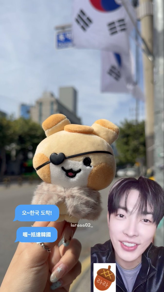A hand holds a plush Shiba Inu with an eye patch in front of a South Korean flag, with a smiling photo of ATEEZ's Hongjoong and a "라라라" (lalala) acorn sticker overlaid, alongside Chinese subtitle bubbles reading "哦~抵達韓國" (Oh~arrived in Korea), referencing the singer's first vlog from Yeongtong on 261005.