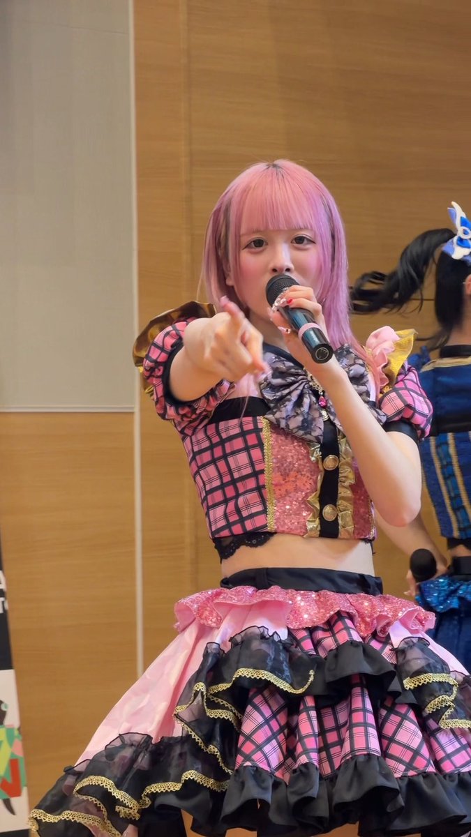 A performer with pink hair wearing a pink and black plaid ruffled outfit points toward the camera while holding a microphone.