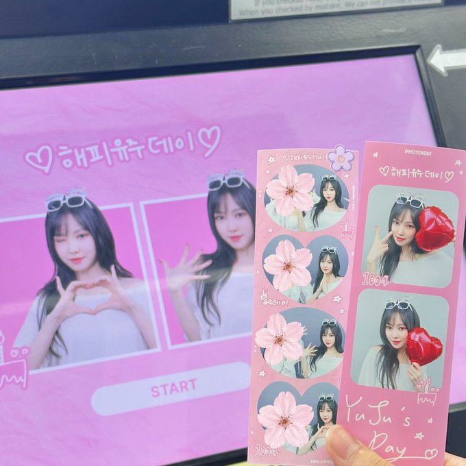 A hand holds a pink photo strip featuring four photos of a young woman with long dark hair, alongside a photo booth screen displaying "해피유주데이" (Happy Yuriday) with pink Korean text.