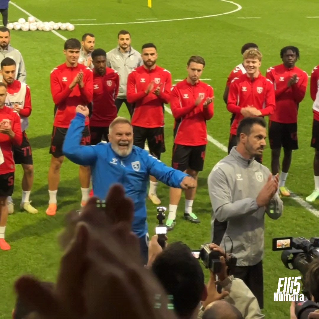 A jubilant older man in a blue tracksuit celebrates with his arms raised in front of a football team wearing red training kits on a pitch at night, with a 'Elif5 Numara' watermark in the corner.