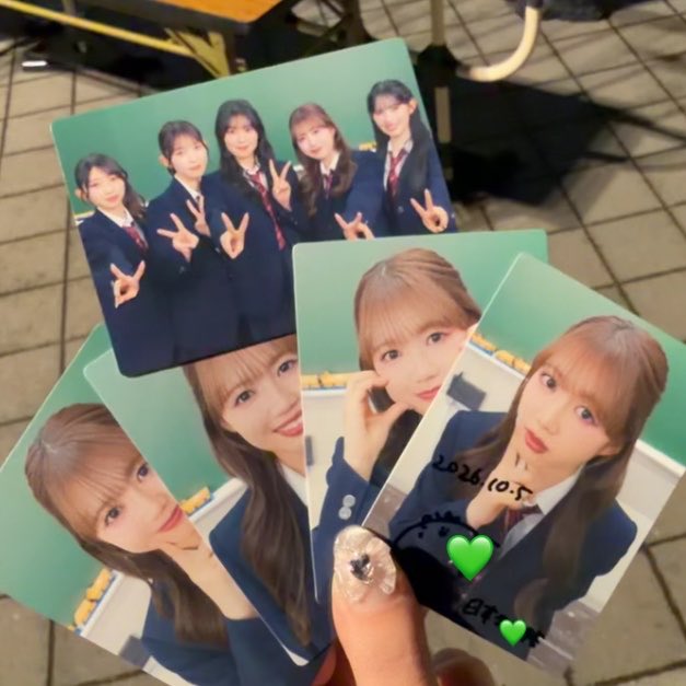 A hand holds several photocards of female idols in dark blue school uniforms; the cards show group shots and solo portraits, with one card marked '2026.10.5' and decorated with green heart stickers.