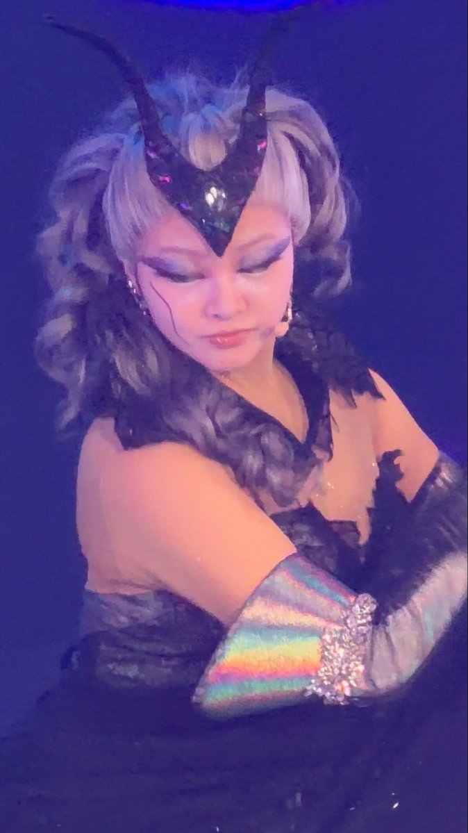 A performer in elaborate stage makeup and costume with dramatic dark eye makeup, a horned headpiece, and rainbow-striped iridescent gloves performs against a blue-lit background.