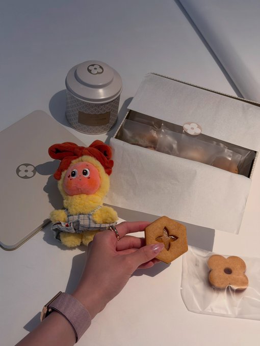 A photo showing a hand holding a star-shaped cookie next to a flower-shaped cookie and a small plush toy, with a tin and box in the background featuring a clover logo.
