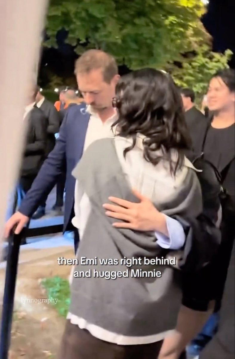 A nighttime scene showing a man in a suit walking while a woman in a gray jacket hugs another woman, with text overlay reading 'then Emi was right behind and hugged Minnie'.