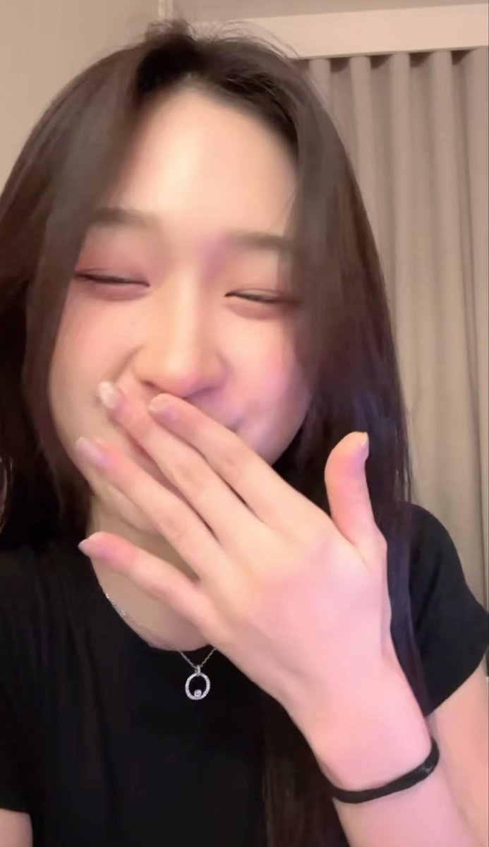 A close-up selfie of a young woman smiling brightly with her eyes closed and her hand covering her mouth.