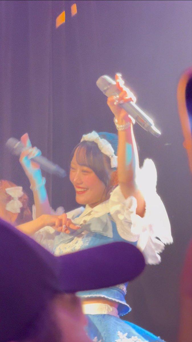 A performer in a blue and white maid-style outfit on stage, smiling and holding a microphone up in one hand.