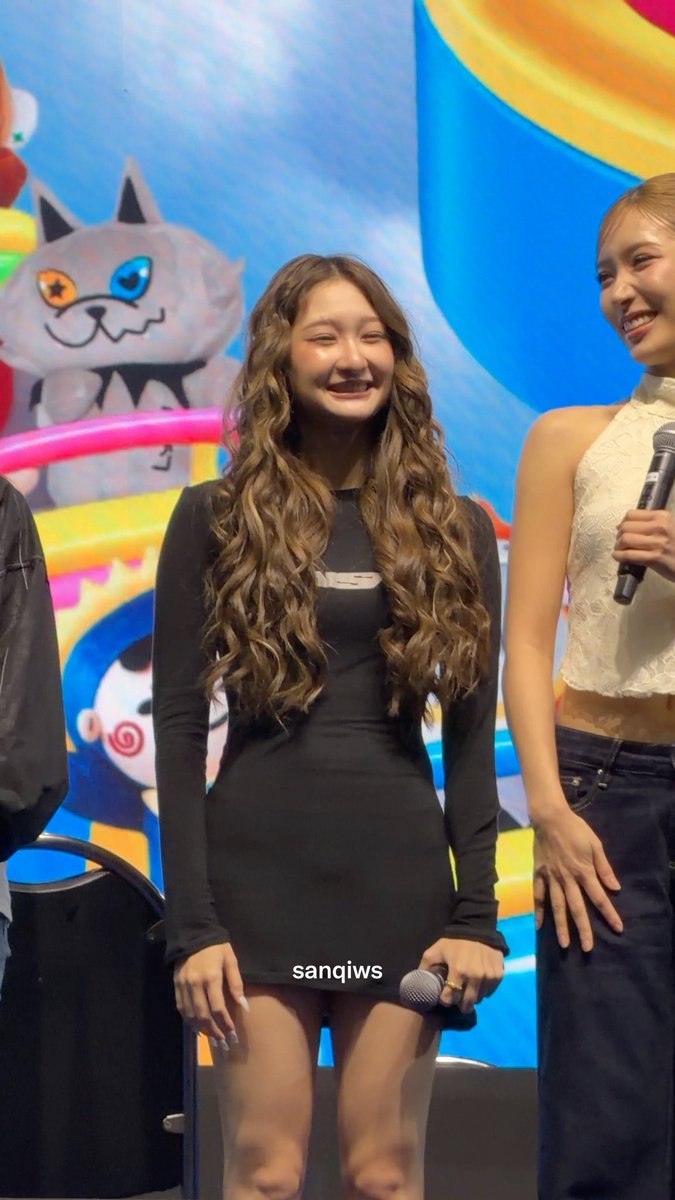 A woman with long curly hair in a black mini dress smiles while holding a microphone on stage, with a 'sanqiws' watermark visible.
