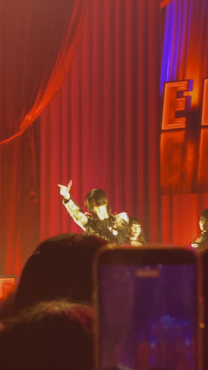 A performer points toward the audience on stage in front of a red curtain, while another member sits behind, as a fan holds up a phone to capture the moment. The image reflects the caption's story of impulsively filming the live performance after being captivated by the vocalist's expression during their first time seeing the act.