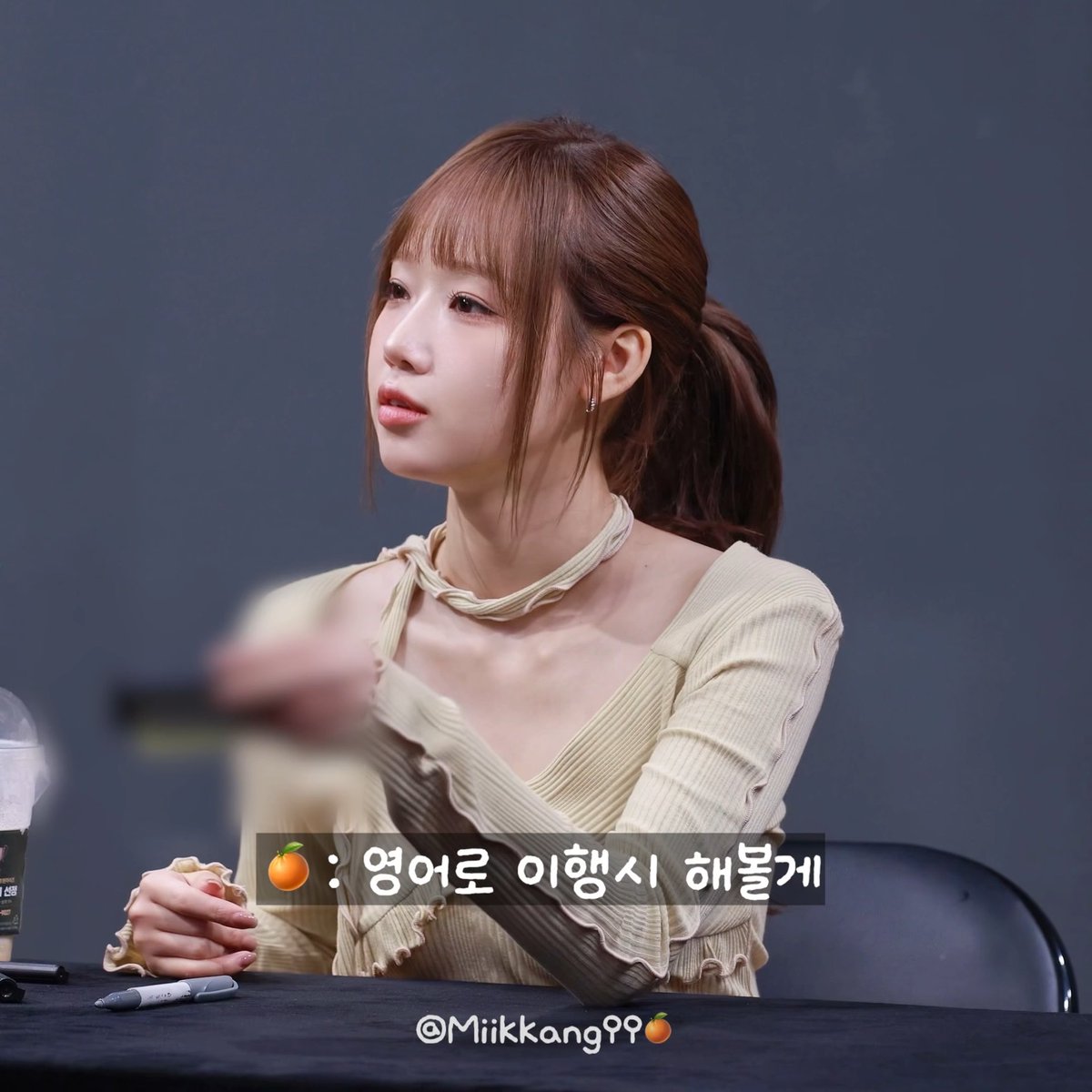 A young woman with reddish-brown hair in a ponytail wearing a beige ribbed top with a matching choker, seated at what appears to be a fan event, with Korean text and the watermark 'Miikkang99'.