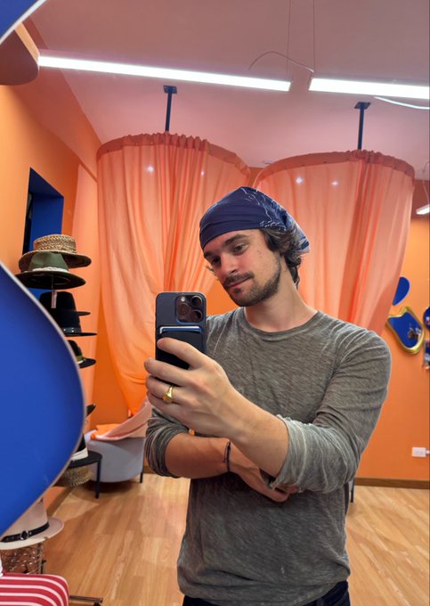 A man with a beard takes a mirror selfie in an orange-walled shop, wearing a blue bandana tied around his head and a gray long-sleeve shirt. The colorful setting, complete with peach curtains and hat displays, complements his playful post asking whether he should bring back the bandana look.