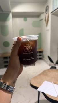 A hand wearing a watch holds a clear plastic cup filled with iced brown coffee from 'Specialty Coffee' inside a room with light green polka-dot walls.