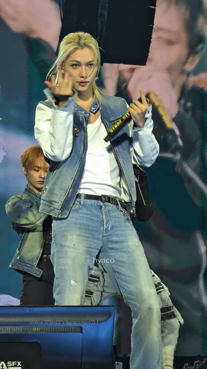 A female K-pop performer with blonde hair performs on stage wearing a denim vest and white top, with backup dancers visible behind her; watermark reads 'hyaco' and '26094 STRAXCITY Buenos Aires D.1'.