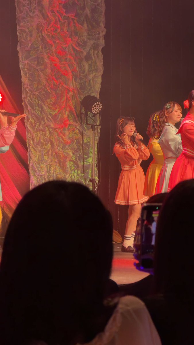 A performer in an orange outfit sings into a microphone on stage alongside others in colorful costumes, with a textured backdrop lit in red and green.