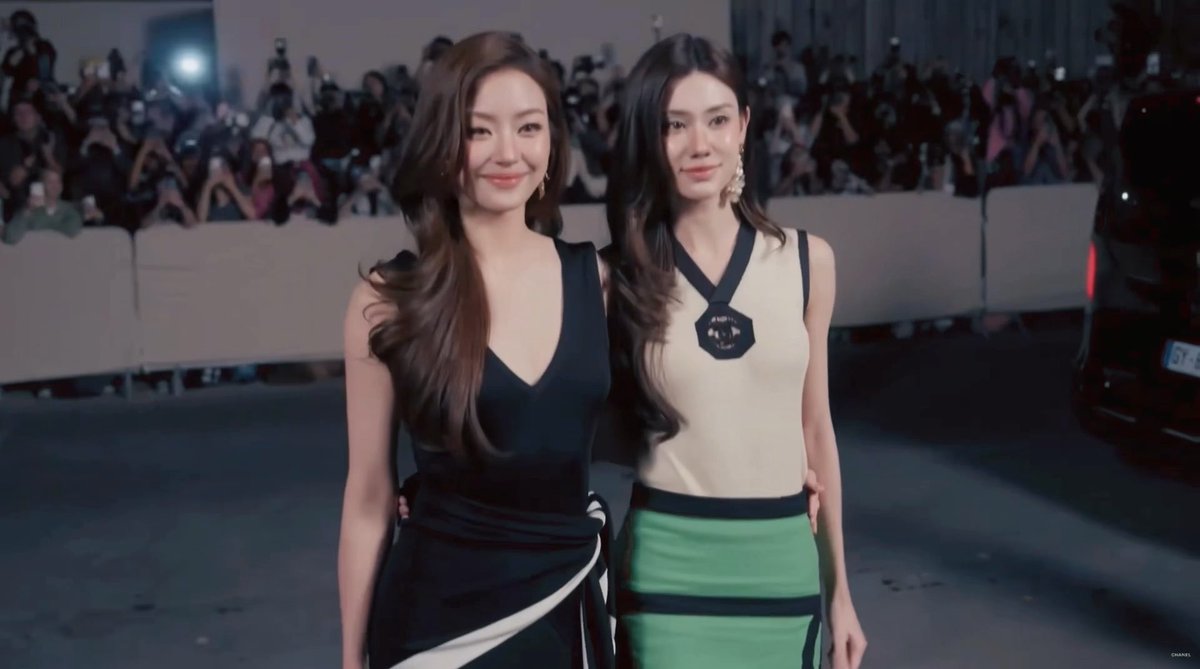 Two women pose together on what appears to be a red carpet event, with one wearing a black dress and the other in a white and green outfit, while photographers crowd the background.