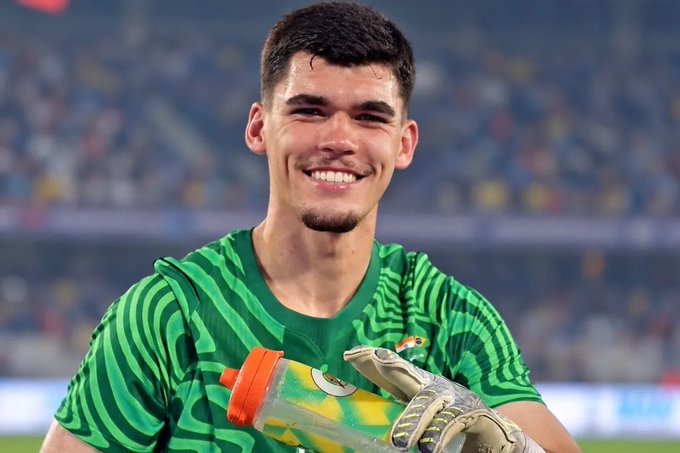 A young goalkeeper wearing a green Brazil national team jersey smiles warmly at the camera while holding a water bottle, his gloved hand visible, capturing the celebratory moment of becoming the 100th goalkeeper to represent the Brazilian national team in history.
